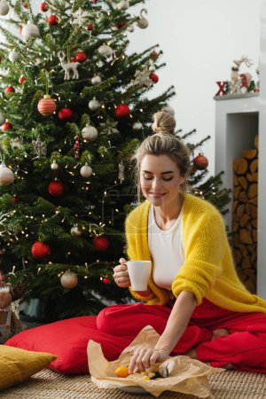 Téléchargez les photos : Une femme heureuse est assise à côté d'une cheminée lumineuse dans un salon confortable, orné d'un arbre de Noël et de décorations festives, profitant d'une tasse de boisson chaude. - en image libre de droit