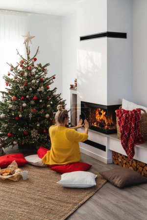 Téléchargez les photos : Jeune femme assise à côté d'une cheminée lumineuse dans un salon confortable orné d'un arbre de Noël et de décorations festives. - en image libre de droit