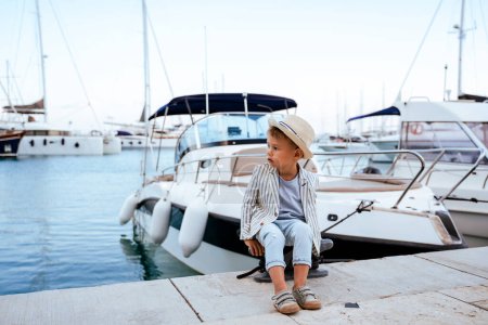 Foto de Lindo chico elegante sentado en el muelle cerca de yate en marina. - Imagen libre de derechos