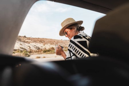Foto de Mujer viajera tomando un descanso con café en su coche en una playa de guijarros. - Imagen libre de derechos