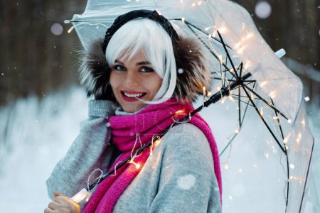 Téléchargez les photos : Femme, ornée d'une perruque blanche, de cache-oreilles thermiques, d'un pull confortable et d'une écharpe rose. Elle tient un parapluie transparent intégré avec des lumières scintillantes, créant une atmosphère hivernale magique. - en image libre de droit