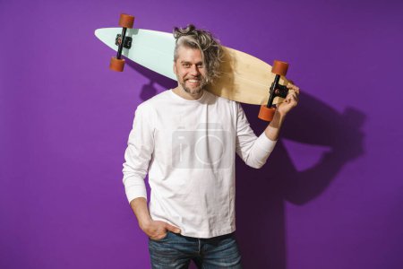 Téléchargez les photos : Portrait de joyeux homme d'âge moyen avec longboard portant sweat-shirt blanc sur fond violet - en image libre de droit