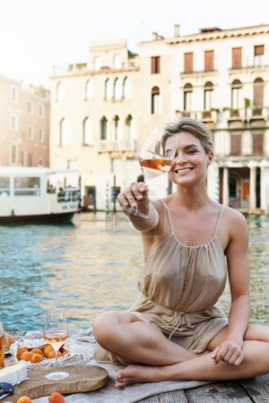 Téléchargez les photos : Belle jeune femme pique-nique pittoresque sur le quai de gondole en bois avec vin rose, fruits et collation sur une jetée en bois à Venise, Italie - en image libre de droit