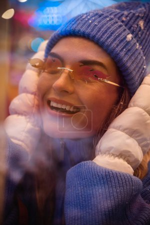 Photo for Cheerful and stylish woman, dressed in warm clothes and wearing sunglasses shaped like fire flames, is having fun in a snowy winter amusement park. - Royalty Free Image