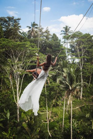 Foto de Mujer joven con vestido blanco balanceándose en columpios de cuerda con hermosa vista sobre terrazas de arroz y palmeras en la isla de Bali - Imagen libre de derechos