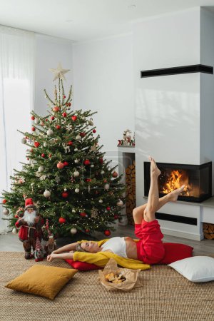 Photo for Young woman lies beside a glowing fireplace in a cozy living room, adorned with a Christmas tree and festive decorations. - Royalty Free Image