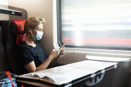 Téléchargez les photos : Portrait de jeune femme portant un masque facial médical à l'aide d'un smartphone pendant son voyage en train - en image libre de droit