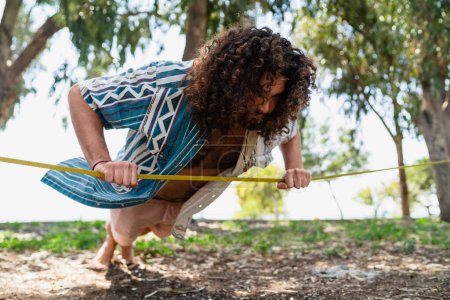 Téléchargez les photos : Jeune homme confiant faisant des pompes et équilibrage sur le slackline dans le parc de la ville pendant la journée d'été - en image libre de droit