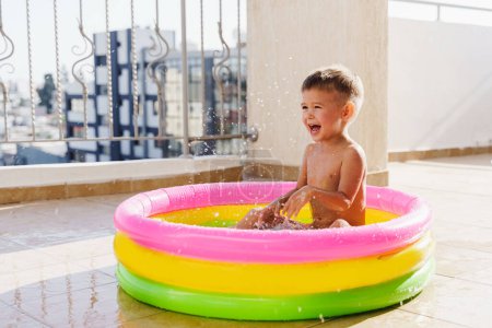 Foto de Niño pequeño juega alegremente en una piscina inflable redonda en el balcón durante un día caluroso de verano. - Imagen libre de derechos