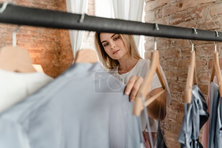 Photo for Young woman is browsing through her various outfits hanging on the clothing rack in her wardrobe - Royalty Free Image