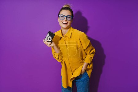 Foto de Retrato de una joven alegre con anteojos que sostiene la cámara de cine vintage sobre fondo púrpura - Imagen libre de derechos
