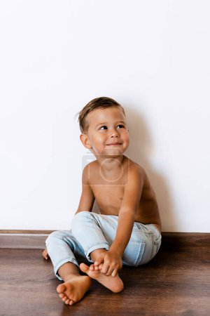 Téléchargez les photos : Portrait de heureux petit garçon joyeux portant un jean posant contre un mur blanc - en image libre de droit