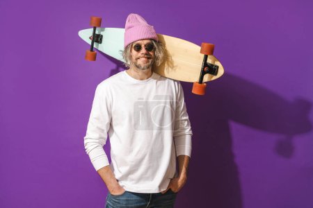 Téléchargez les photos : Portrait de joyeux homme d'âge moyen avec longboard portant sweat-shirt blanc sur fond violet - en image libre de droit