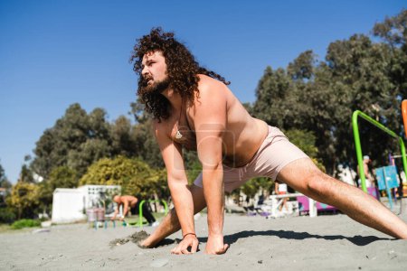 Téléchargez les photos : Jeune homme bouclé et barbu pendant son entraînement d'étirement sur la plage - en image libre de droit