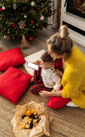 Photo for Woman and her little son look at a smartphone with a blank screen in a cozy living room adorned with a Christmas tree and festive decorations. - Royalty Free Image