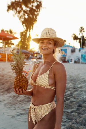 Foto de Joven mujer feliz disfrutando de cóctel en piña durante el atardecer en la playa. - Imagen libre de derechos
