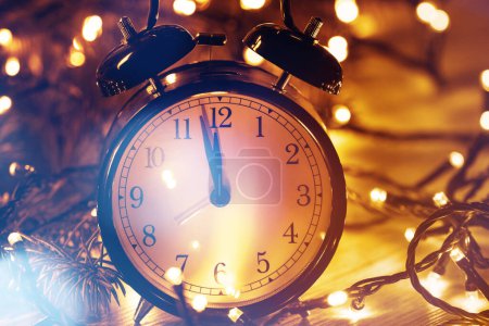 Foto de Reloj despertador cerca de medianoche, rodeado de radiantes luces de Navidad y ramitas de abeto fresco en una superficie de madera rústica. - Imagen libre de derechos