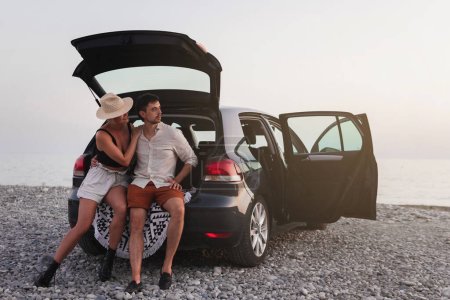 Téléchargez les photos : Couple de voyageurs debout à côté de leur voiture sur une plage de galets pendant leur voyage en voiture. - en image libre de droit