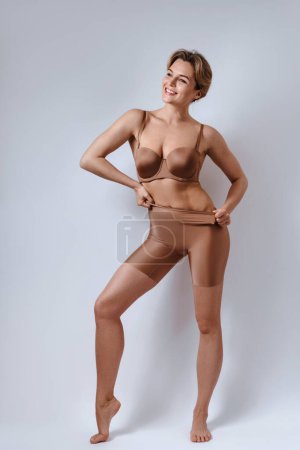 Téléchargez les photos : Portrait de jeune femme séduisante portant une lingerie marron avec des culottes taille haute sur fond gris. - en image libre de droit