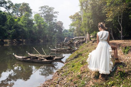 Photo for Beautiful young woman wearing white robe dress posing near river with old wooden boats - Royalty Free Image