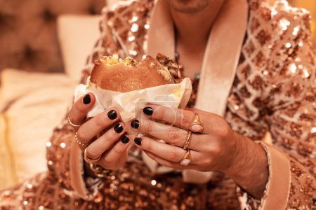 Téléchargez les photos : Homme riche dans un costume doré chatoyant portant des anneaux d'or et mangeant un hamburger. - en image libre de droit