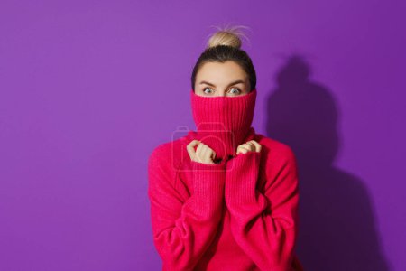 Foto de Retrato de mujer joven está escondiendo su cara dentro de un suéter de cuello de polo cálido y acogedor sobre fondo púrpura - Imagen libre de derechos