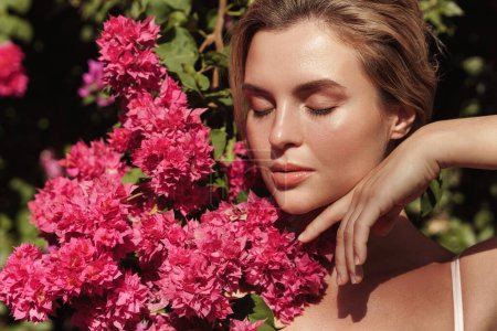 Téléchargez les photos : Belle femme blonde à la peau lisse et au maquillage naturel, posant sur fond de buisson aux délicates fleurs roses. - en image libre de droit