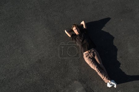 Téléchargez les photos : Vue du dessus du jeune homme adulte insouciant couché sur l'asphalte - en image libre de droit