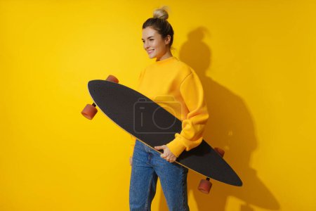 Téléchargez les photos : Portrait de jeune fille gaie avec longboard sur fond jaune - en image libre de droit