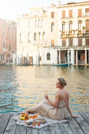 Photo for Lovely young woman picturesque picnic on the wooden gondola dock  with rose wine, fruits and snack on wooden pier in the Venice, Italy - Royalty Free Image