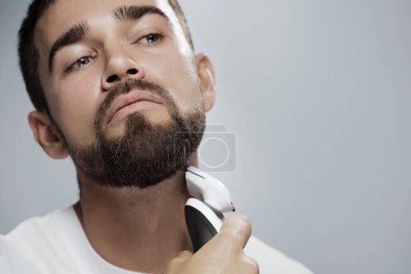 Foto de Joven hombre guapo está utilizando trimmer eléctrico para afeitarse la barba contra el fondo gris - Imagen libre de derechos