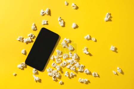 Téléchargez les photos : Parsemé de délicieux pop-corn et smartphone avec écran blanc sur fond jaune - en image libre de droit