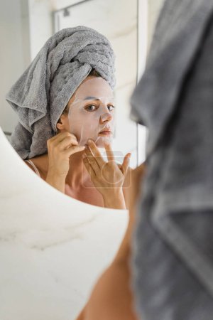 Photo for Beautiful woman in bathroom with applied sheet mask on her face looking in the mirror - Royalty Free Image
