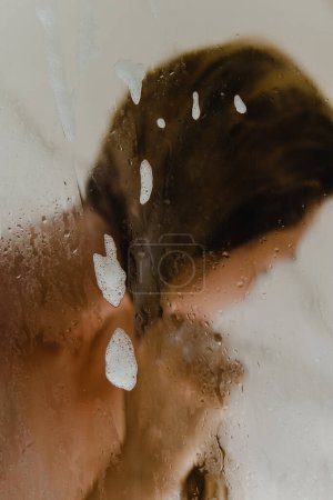 Téléchargez les photos : Jeune femme se lave les cheveux avec un shampooing tout en prenant une douche. Focus sur le verre mouillé. - en image libre de droit