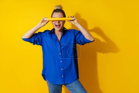 Téléchargez les photos : Joyeux jeune femme portant chemise bleue tenant crayon géant sur fond jaune - en image libre de droit