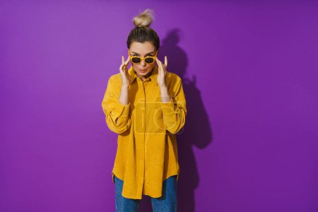 Photo for Portrait of young  girl wearing yellow shirt and sunglasses making silly face expression against purple background - Royalty Free Image