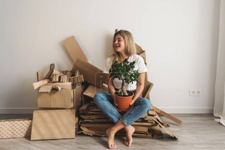 Photo for Young woman with small ficus plant and collected cardboard waste at home. Concepts of Paper Recycling and Waste Sorting and Saving Trees. - Royalty Free Image