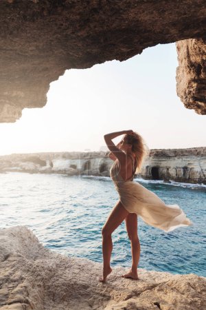 Téléchargez les photos : Femme en robe de soie fluide sur une falaise au coucher du soleil. - en image libre de droit