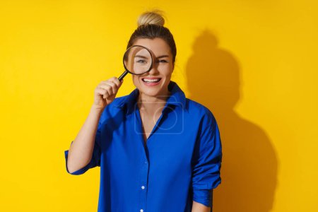 Photo for Curious woman is peering through magnifying glass with great interest, examining something closely on yellow background. - Royalty Free Image