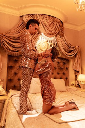 Téléchargez les photos : Le jeune couple riche est vêtu de tenues dorées rayonnantes ornées de broderies à paillettes, situées dans une suite luxueuse. - en image libre de droit