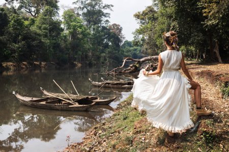 Foto de Hermosa mujer joven con vestido de bata blanca posando cerca del río con viejos barcos de madera - Imagen libre de derechos