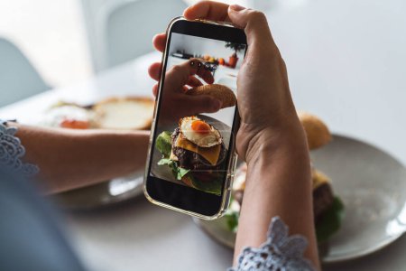 Foto de Manos femeninas usando un smartphone para fotografiar una deliciosa hamburguesa casera con queso y un huevo frito encima. - Imagen libre de derechos