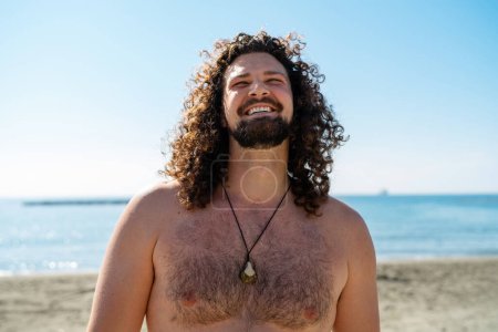 Téléchargez les photos : Portrait de l'homme bouclé joyeux relaxant sur la plage pendant la journée ensoleillée d'été - en image libre de droit