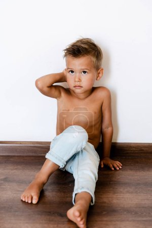 Téléchargez les photos : Mignon petit garçon portant un jean bleu posant contre un mur blanc comme un mannequin. - en image libre de droit