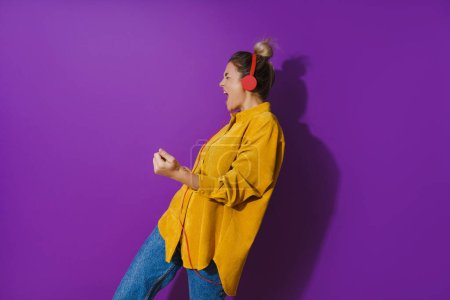 Foto de Retrato de una joven alegre con camisa amarilla escuchando música usando auriculares rojos y bailando sobre fondo púrpura - Imagen libre de derechos