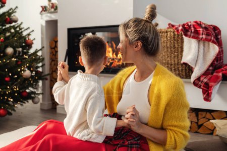 Téléchargez les photos : Jeune femme et son petit fils dansent à côté d'une cheminée dans un salon confortable orné d'un arbre de Noël et de décorations festives. - en image libre de droit
