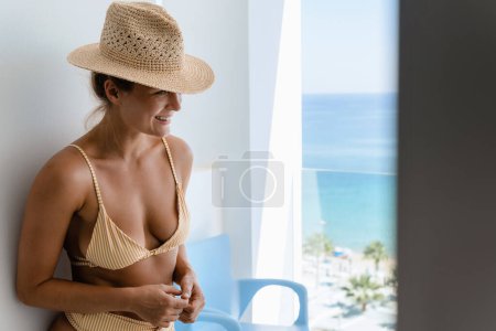 Téléchargez les photos : Femme joyeuse portant un chapeau de paille et un bikini sur le balcon de l'hôtel ou de l'appartement en bord de mer. - en image libre de droit