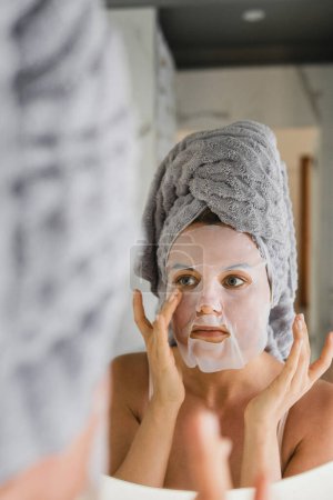 Foto de Hermosa mujer en el baño con máscara de hoja aplicada en su cara mirando en el espejo - Imagen libre de derechos
