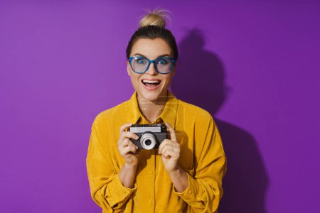 Foto de Retrato de una joven alegre con anteojos que sostiene la cámara de cine vintage sobre fondo púrpura - Imagen libre de derechos