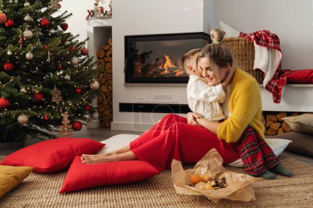 Photo for A young woman and her little son are  beside a glowing fireplace in a cozy living room adorned with a Christmas tree and festive decorations. - Royalty Free Image